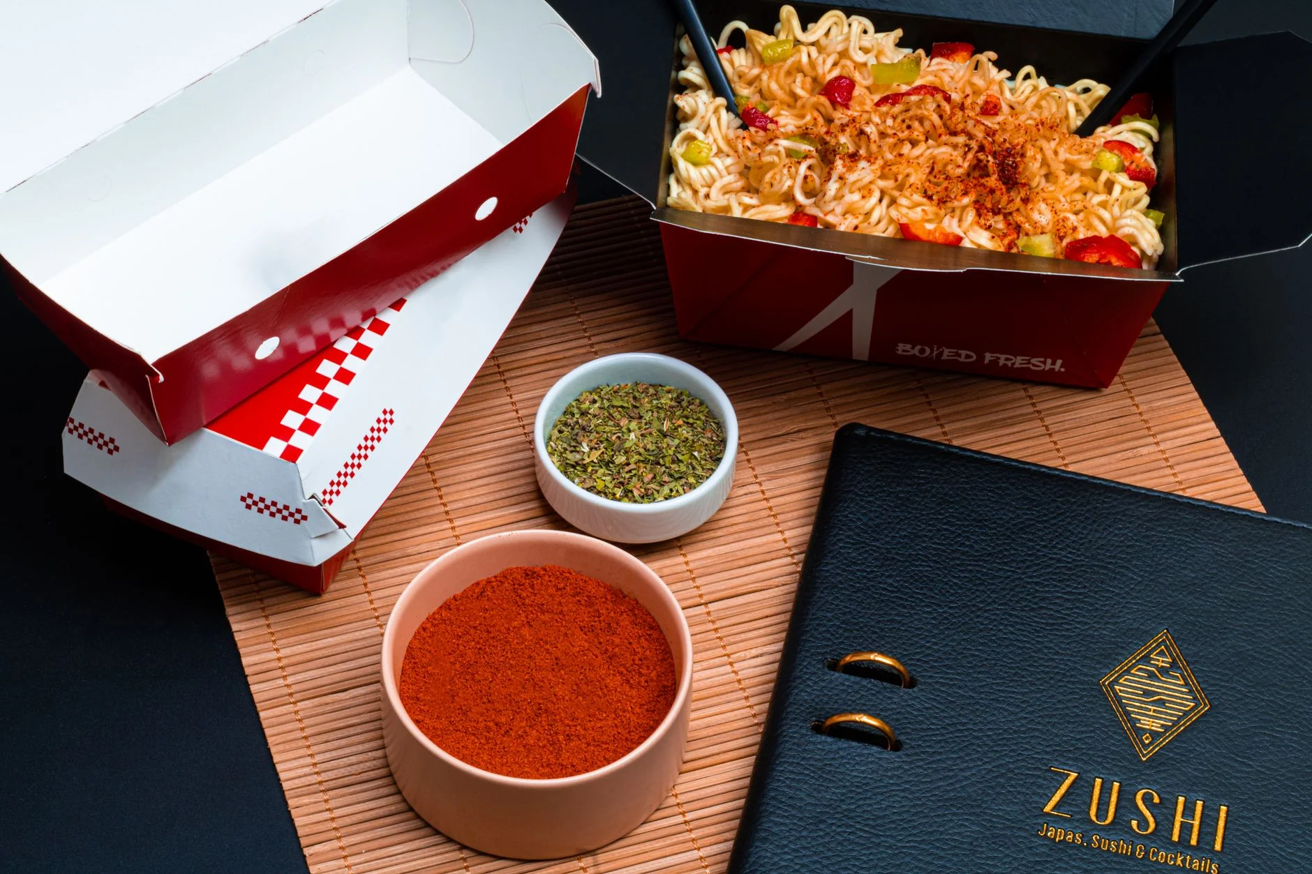 High-angle view of a noodle box, spices, and menu on a bamboo mat backdrop.